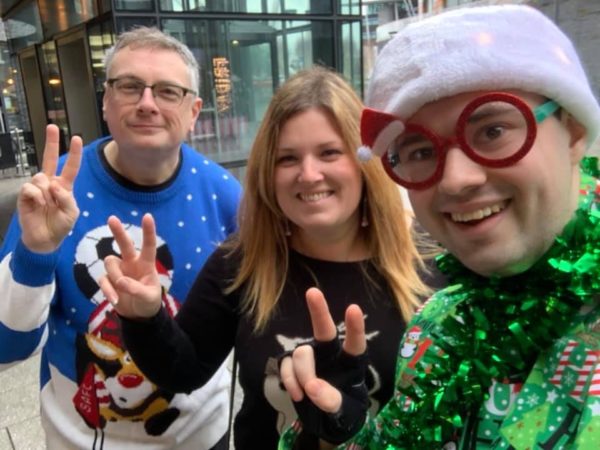 Gerraint poses with 2 colleagues. They are smiling and making the peace sign with their fingers. They are dressed in Christmas jumpers. Gerraint also wear Christmas glasses, hat and green tinsel.
