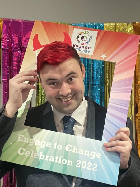 a close up of a young man in a black suit and with bright red hair with his head sticking through the centre of a brightly coloured board that says Engage to Change celebration
