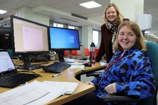 a woman in a blue and black shirt is sitting at a computer with another woman wearing a black suit standing next to her